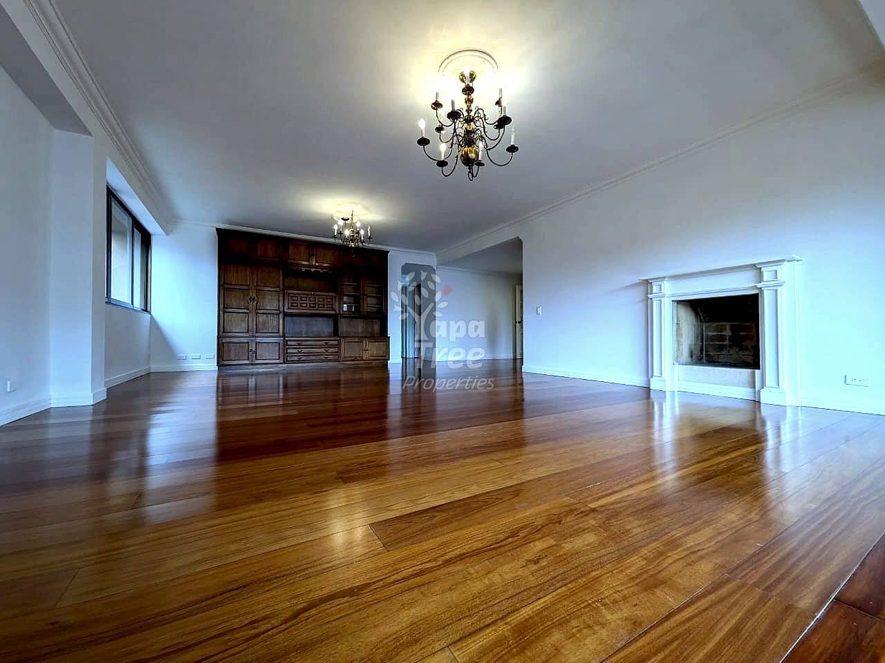 Empty living room with polished wooden flooring, ornate ceiling chandelier, built-in wood cabinetry, fireplace, and large window with natural light
