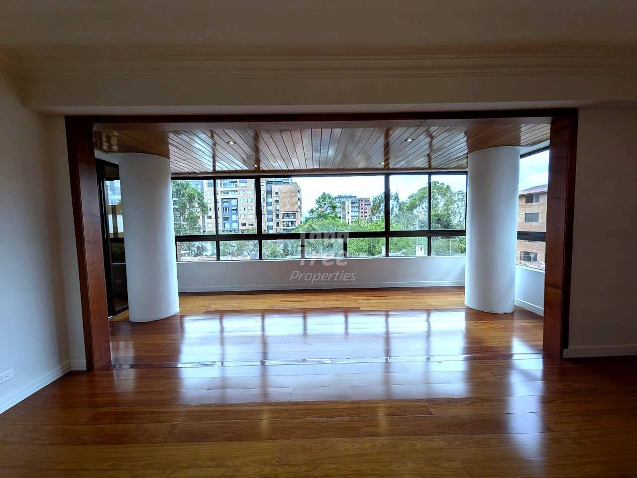 Open living room with wooden floor, two white cylindrical columns, large windows with wood-slatted blinds overlooking urban buildings and trees, natural daylight.