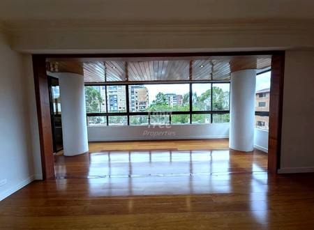 Open living room with wooden floor, two white cylindrical columns, large windows with wood-slatted blinds overlooking urban buildings and trees, natural daylight.