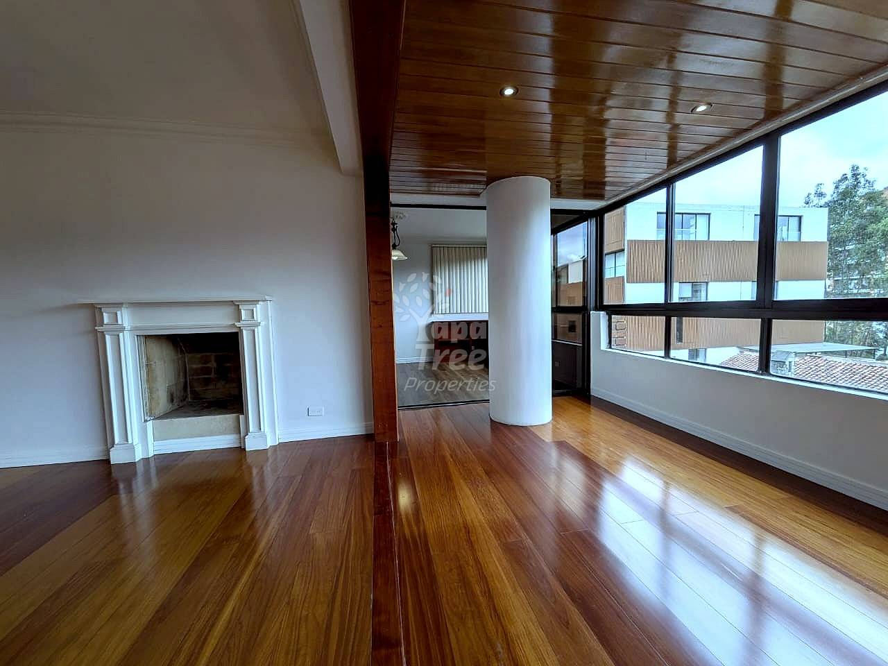 Open living room with wood flooring, white fireplace, central support column, wood ceiling with recessed lighting, and large windows overlooking neighboring buildings.