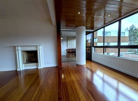 Open living room with wood flooring, white fireplace, central support column, wood ceiling with recessed lighting, and large windows overlooking neighboring buildings.