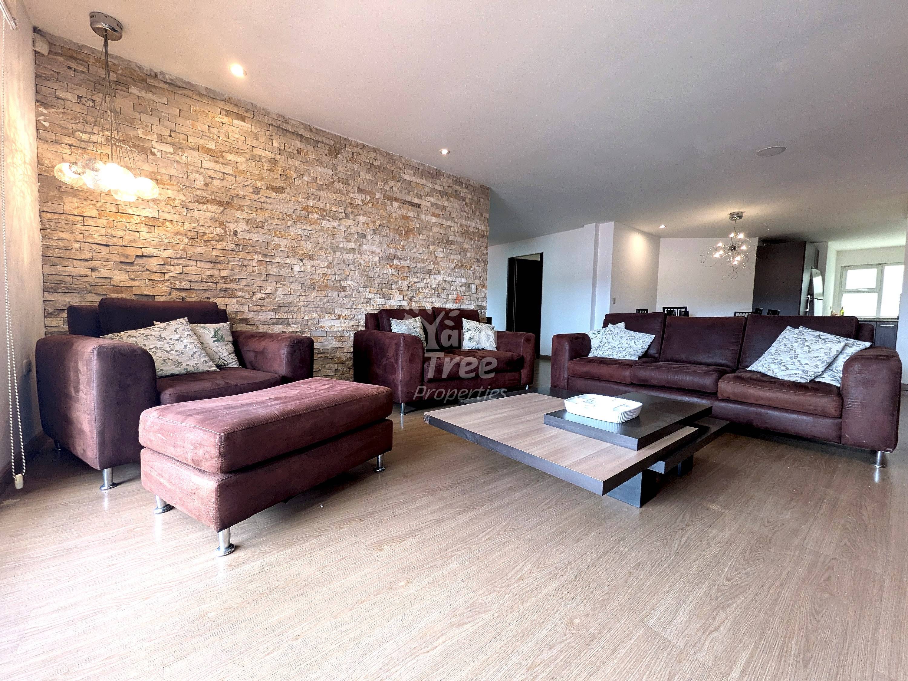 Living room with burgundy upholstered sofas, wood coffee table, light wood flooring, and textured brick accent wall with pendant lighting.