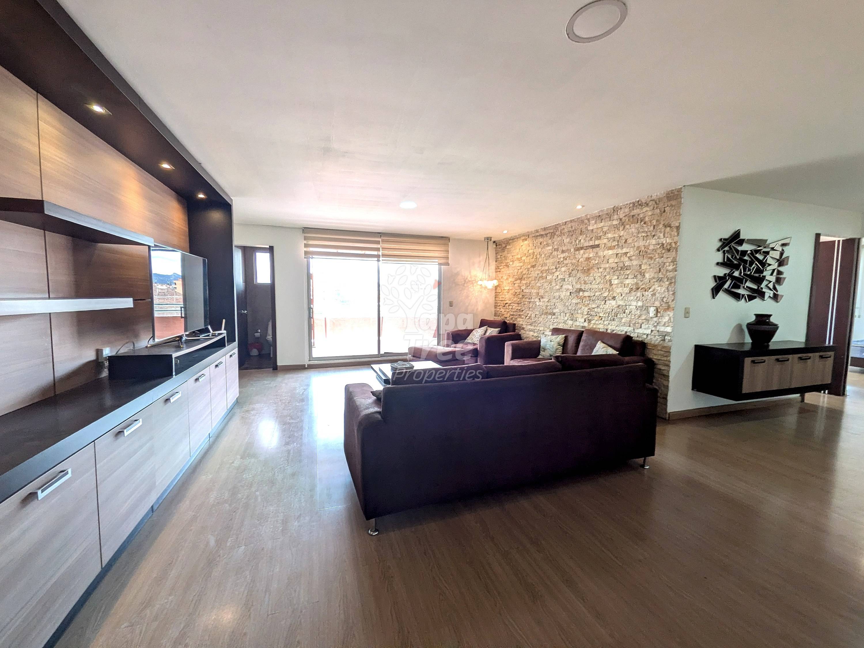 Open-plan living room and kitchen with dark leather sofa, wooden flooring, stone accent wall, built-in cabinetry, and glass sliding doors to balcony.