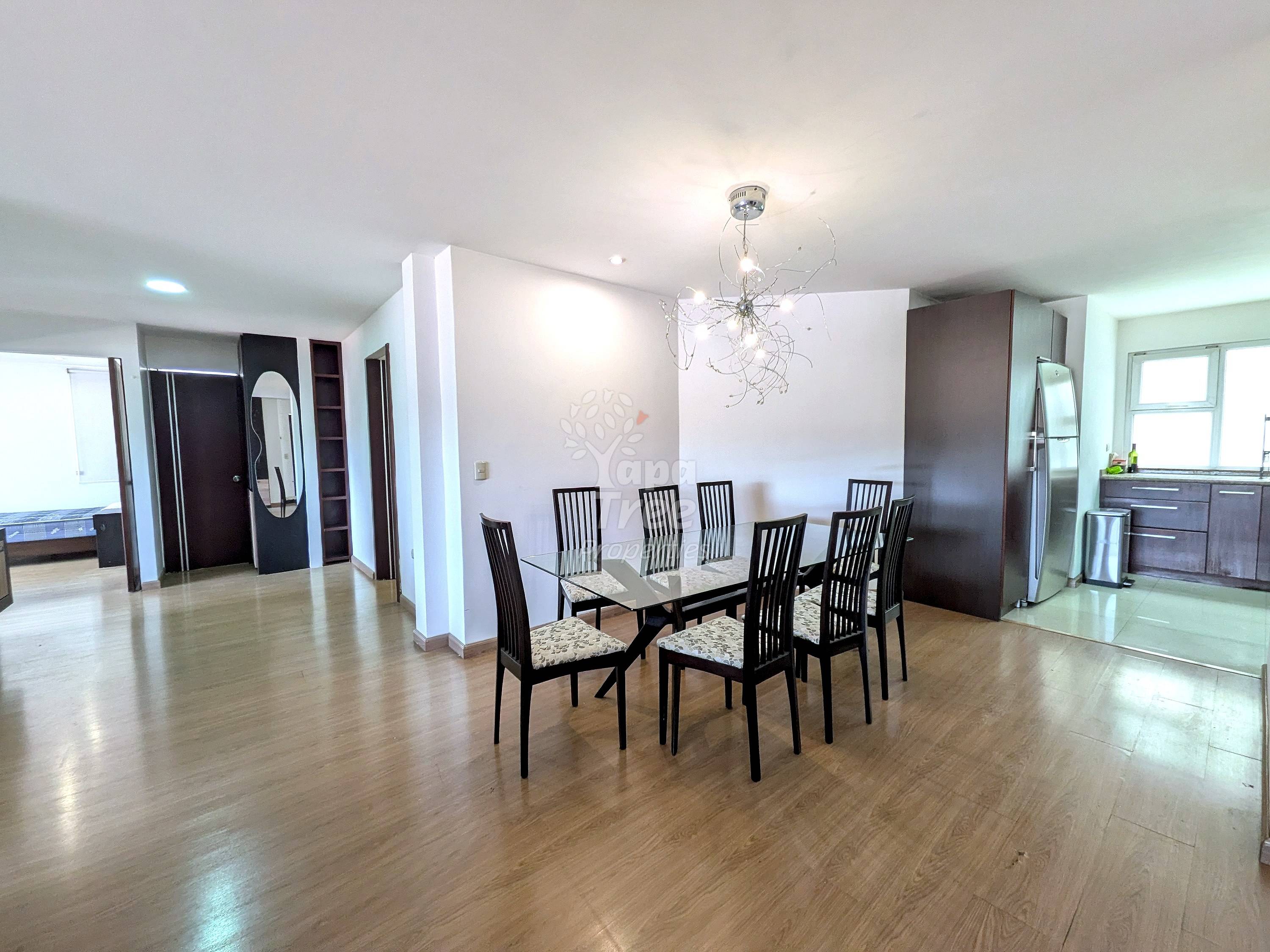 Open-plan dining and living area with wood flooring, dining table with six black chairs, modern pendant light fixture, and kitchen visible in background.
