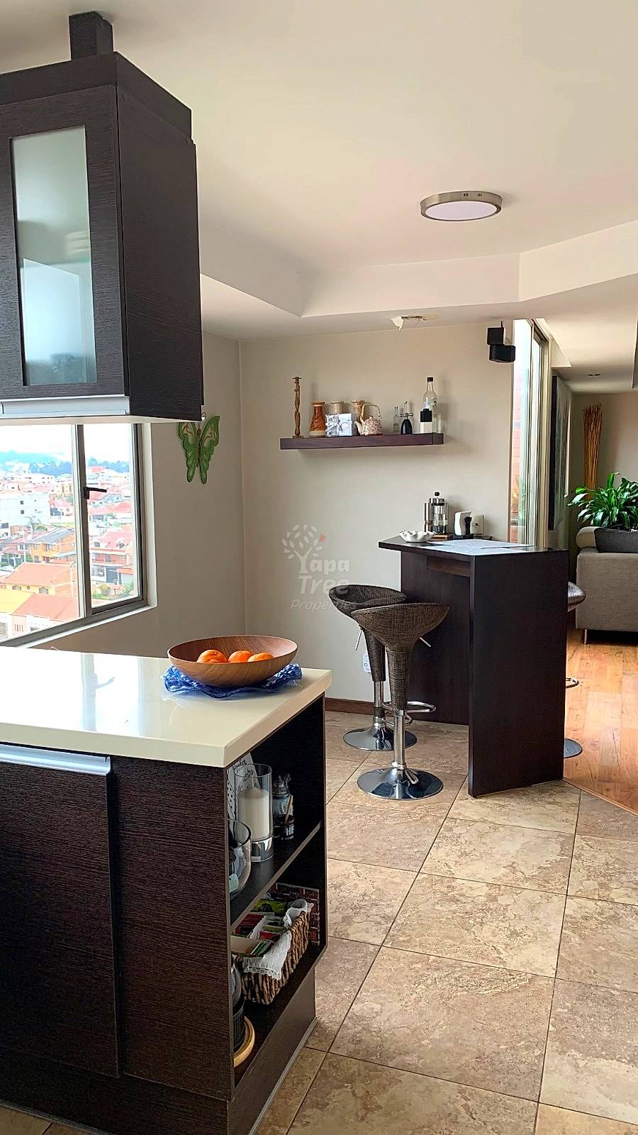 Modern kitchen with dark wood cabinetry, white countertops, tile flooring, bar seating, and large window overlooking Cuenca cityscape.