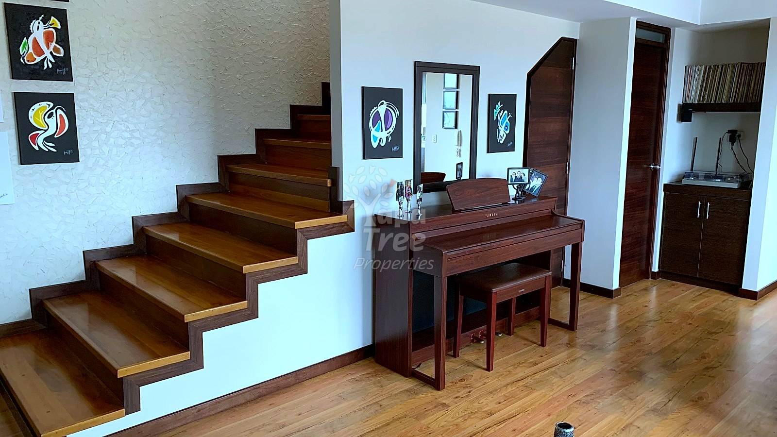 Open living area with wooden staircase, dark wood piano, wooden flooring, and framed artwork on cream walls.