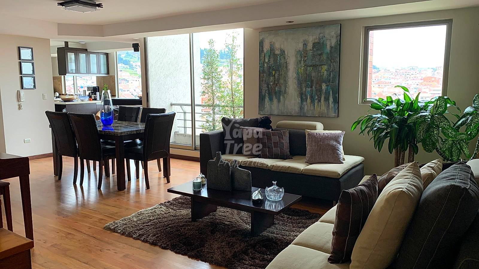 Open-plan living area with wooden flooring, black dining table with chairs, beige and black sofas, city views through large windows, and potted plants.