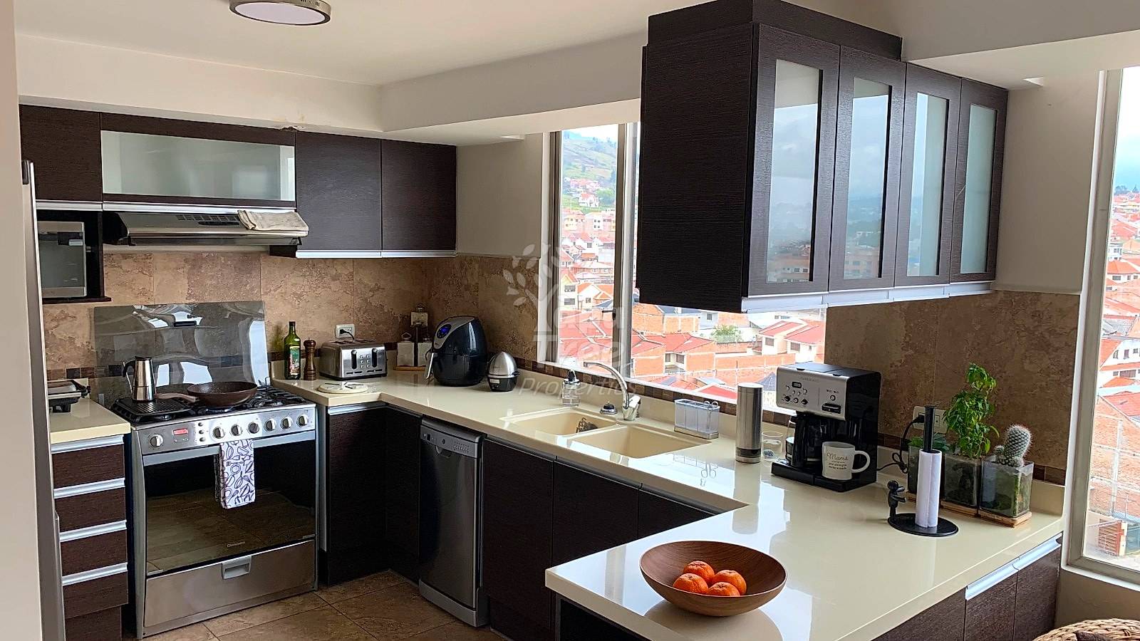 Modern cocina with black cabinets, stainless steel appliances, white countertops, and large windows overlooking Cuenca cityscape with terracotta roofs.