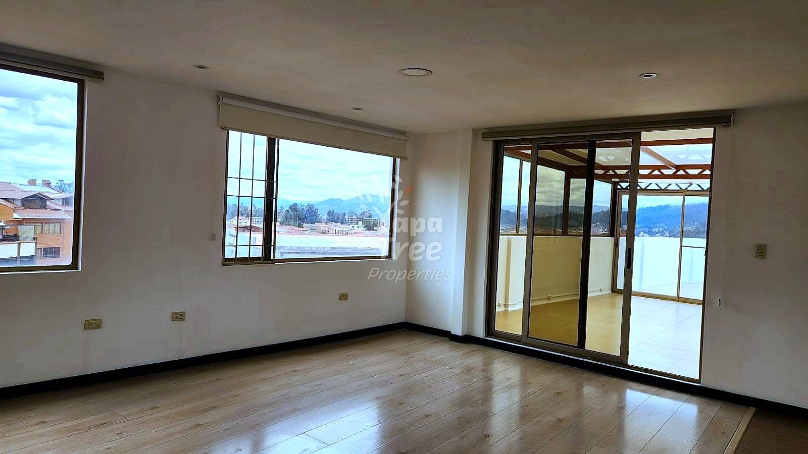 Empty living room with wooden floor, beige walls, multiple windows with mountain views, and glass door leading to terrace.