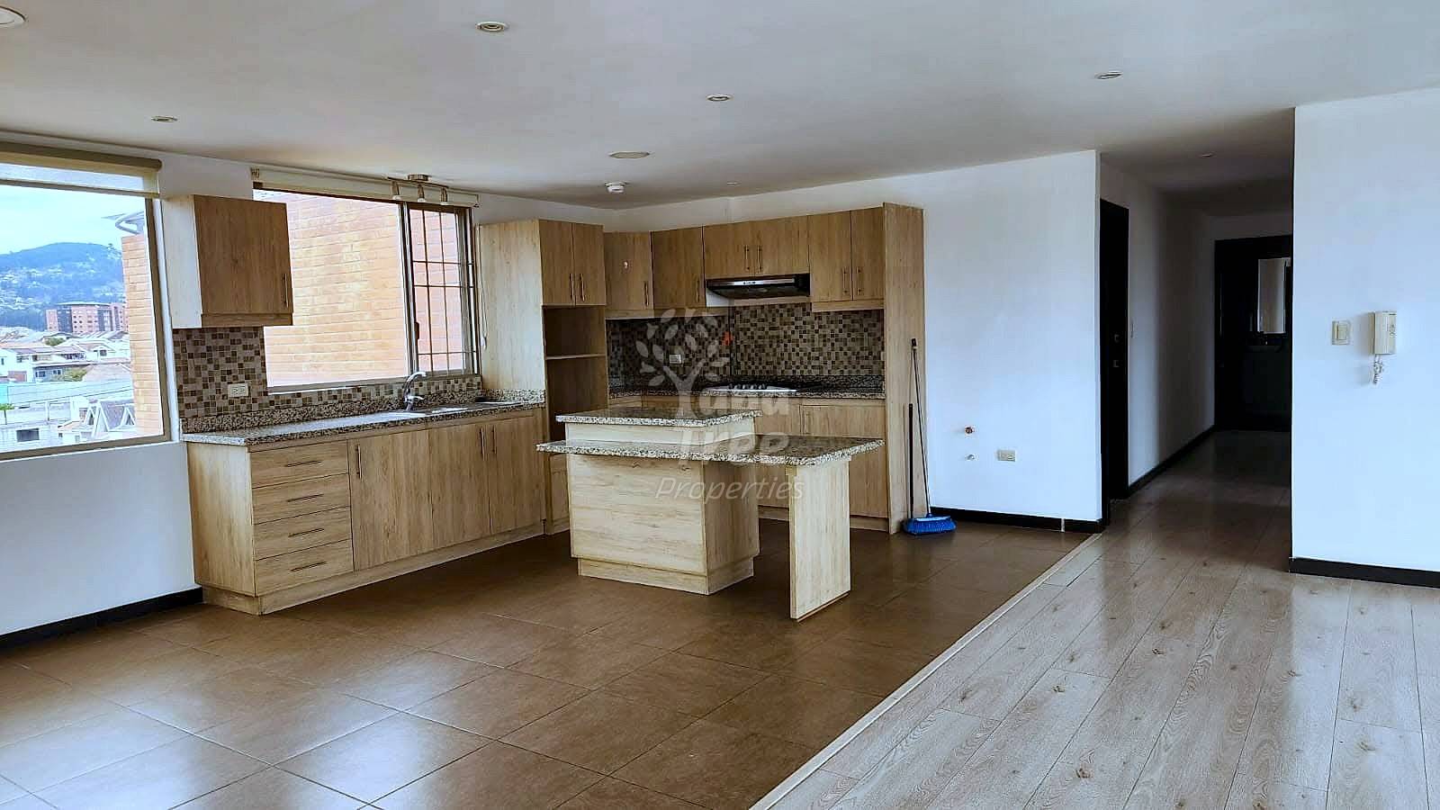Open-concept cocina with wood cabinetry, mosaic tile backsplash, island, and brown tile flooring; living area with wood-look flooring and window overlooking Cuenca cityscape.
