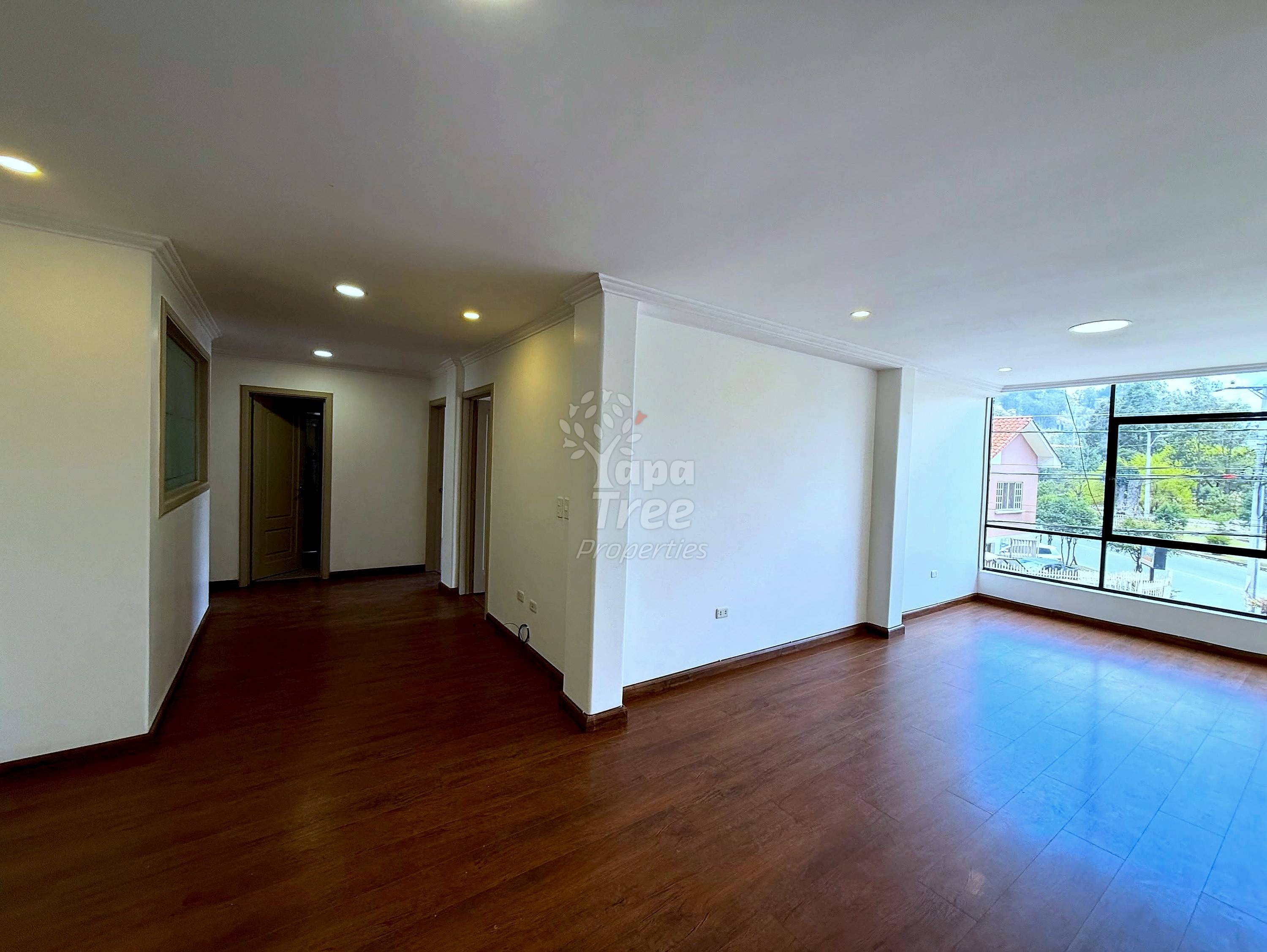 Open living area with wooden floors, white walls, recessed ceiling lights, and large windows overlooking neighborhood buildings and trees.