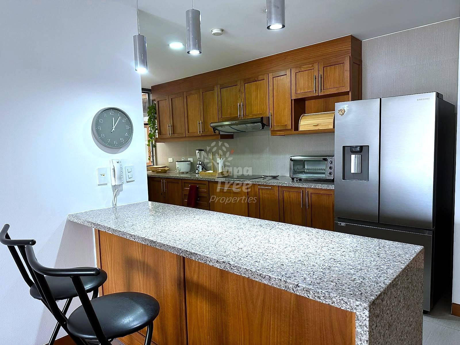 Modern cocina with wood cabinetry, granite countertops, stainless steel refrigerator, pendant lighting, and island seating with black chairs.