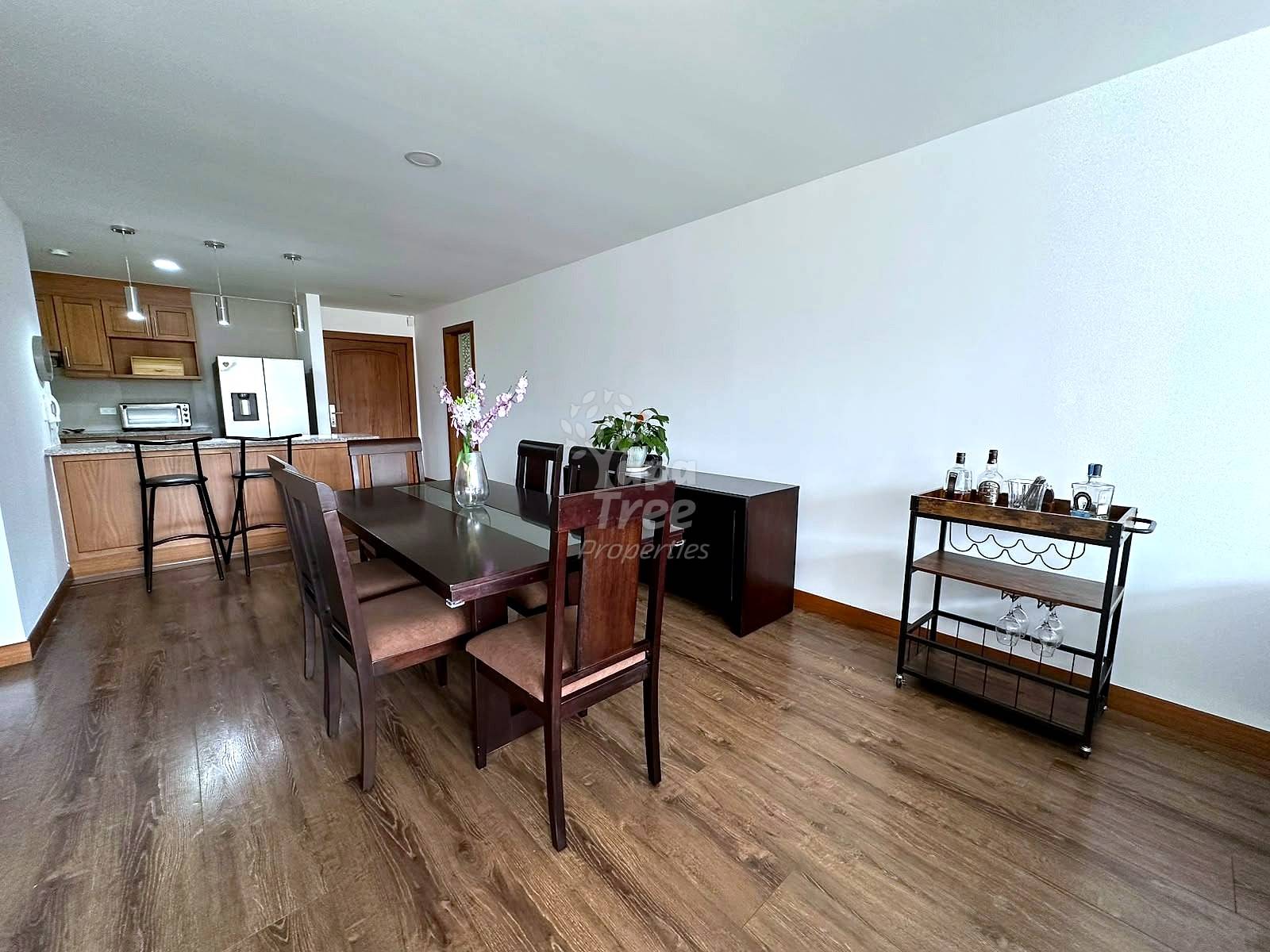 Open-plan dining and kitchen area with wood table, chairs, wooden cabinetry, white appliances, and wood-grain flooring under recessed lighting.