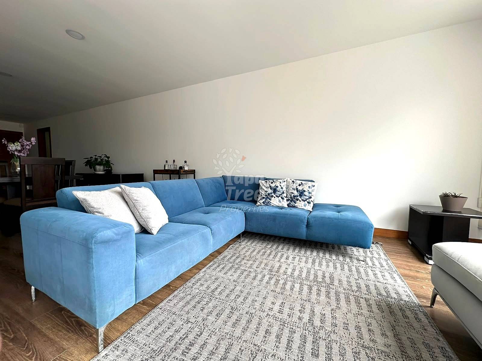 Living room with blue sectional sofa, patterned area rug, wooden flooring, white walls, and recessed ceiling lights.