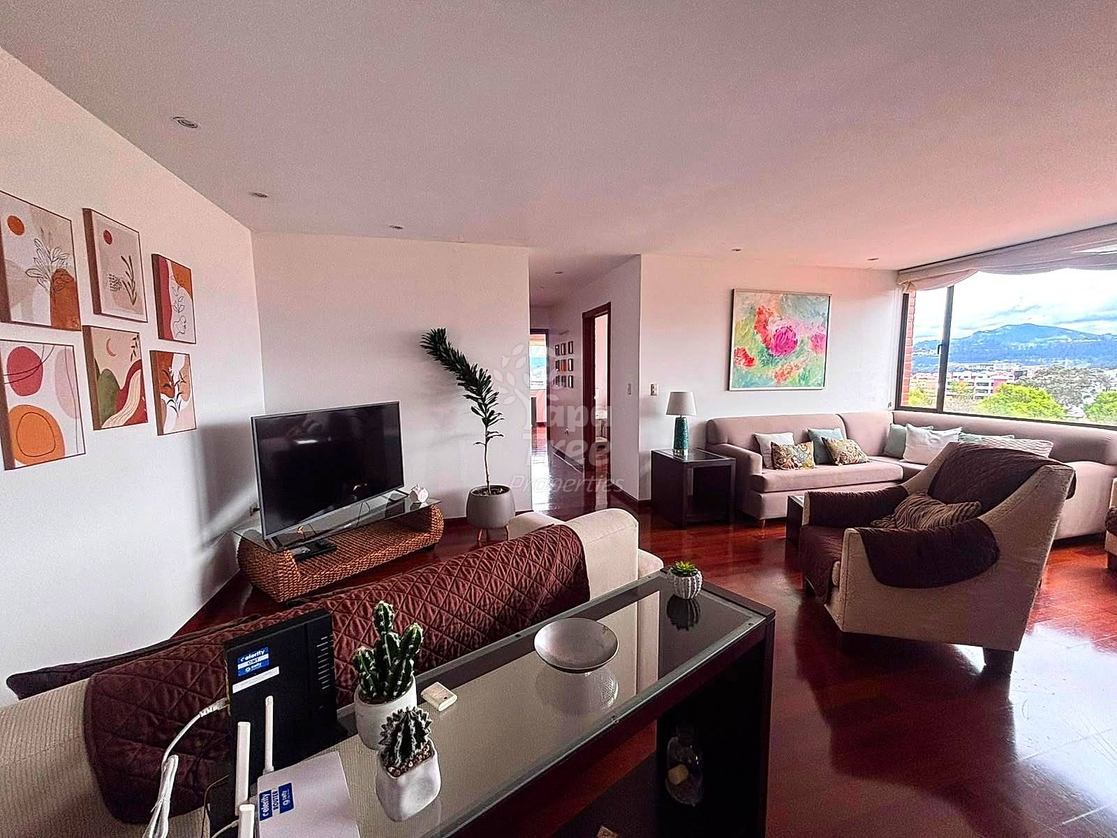 Open-plan living room with terracotta tile floors, beige walls, gray sectional sofa, TV on woven stand, abstract wall art, and large windows overlooking Cuenca cityscape and mountains.