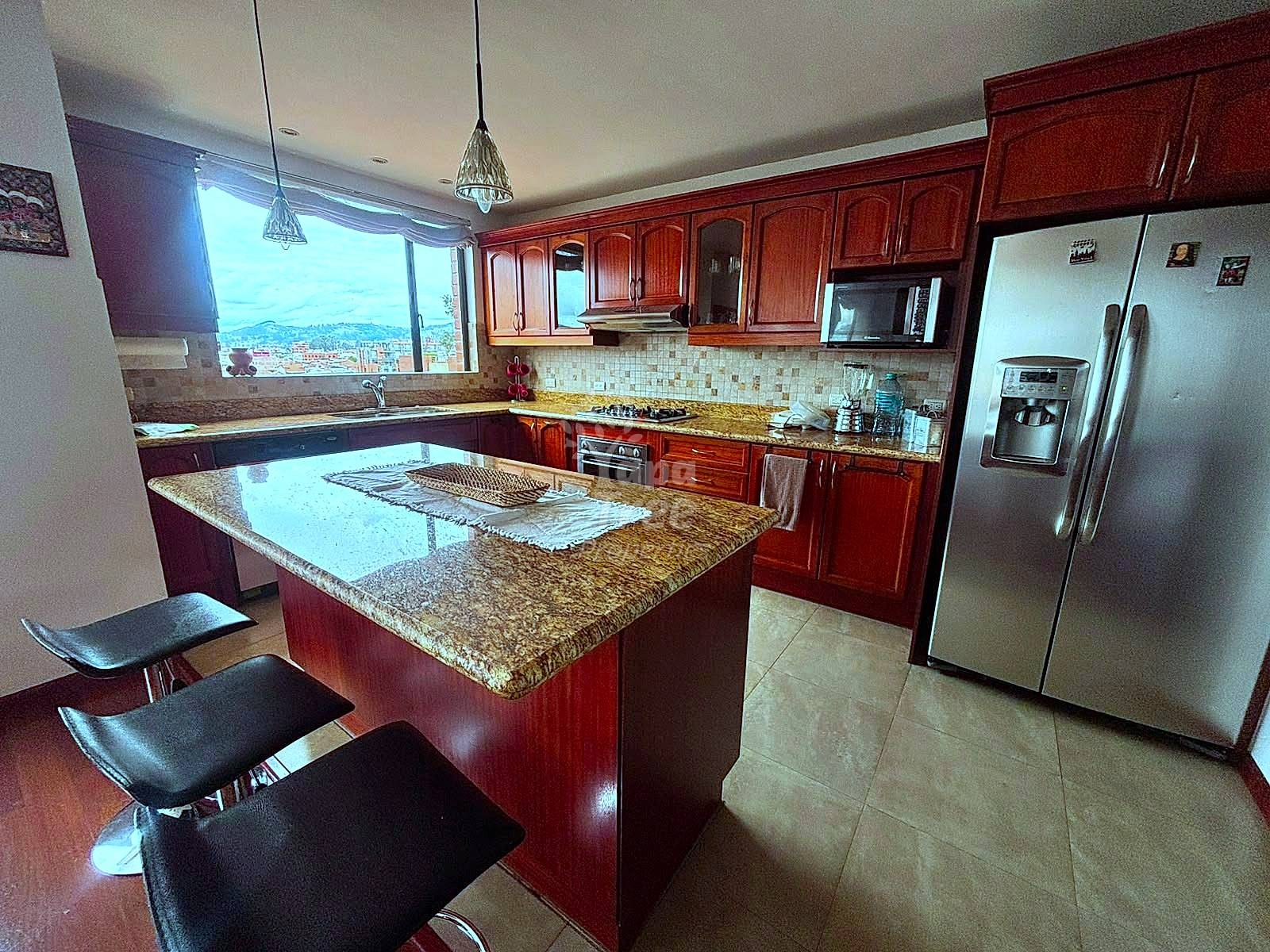 Kitchen with red wood cabinetry, granite island with black bar stools, stainless steel appliances, and window overlooking Cuenca city.