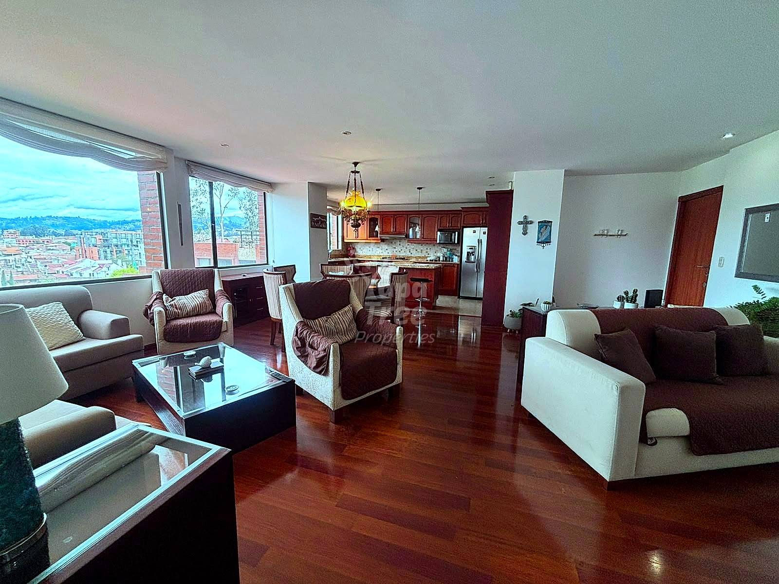 Open-plan living room with burgundy wood flooring, multiple upholstered chairs, white and brown sofas, kitchen with dark cabinetry visible in background, large windows with city views.