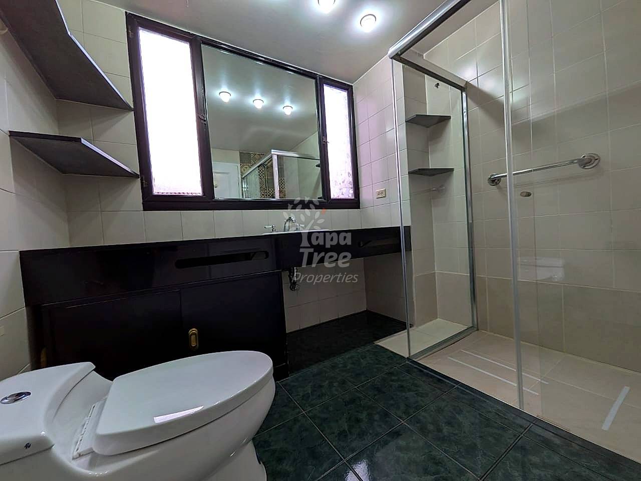 Modern bathroom with white tile walls, black vanity cabinet, illuminated mirror, white toilet, and glass shower enclosure with dark floor tiles.