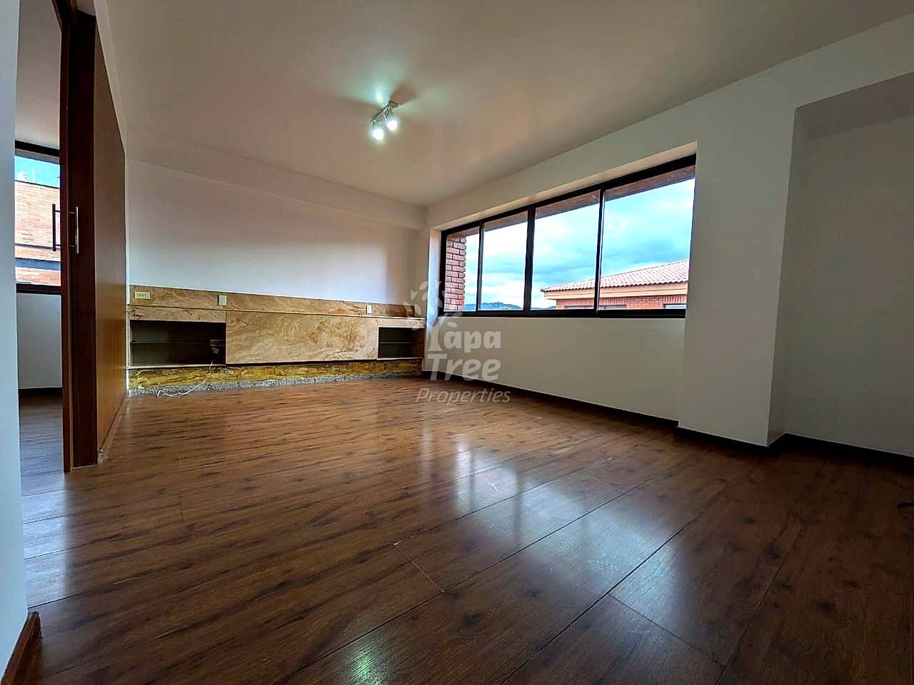 Empty living room with wooden flooring, white walls, built-in stone accent wall with shelving, ceiling lights, and large windows overlooking neighborhood rooftops.