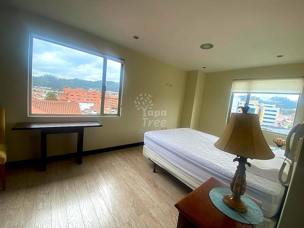 A bedroom features light wood laminate floors, a mattress on a white base, and windows overlooking the city.