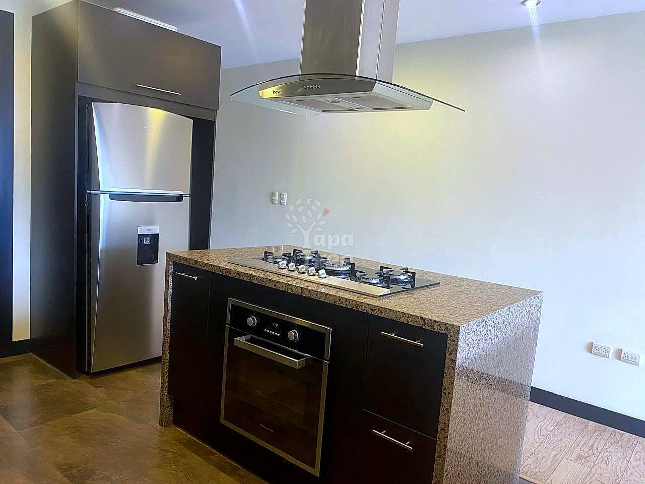 A modern kitchen with dark wood cabinets, a granite island, built-in stove and oven, and a stainless steel refrigerator.