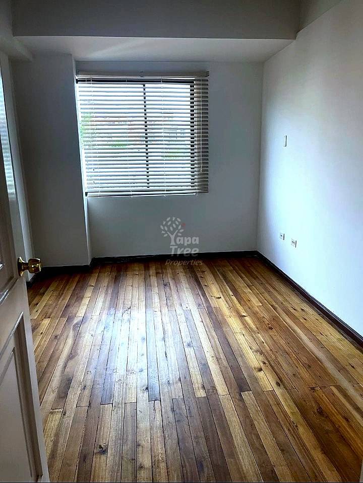 Empty bedroom with wooden floor, white walls, window with horizontal blinds, and electrical outlets.