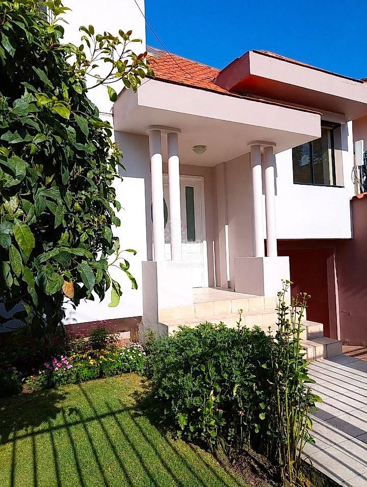Modern white house exterior with columned entrance, terracotta tile roof, manicured front garden with green lawn and flowering plants.