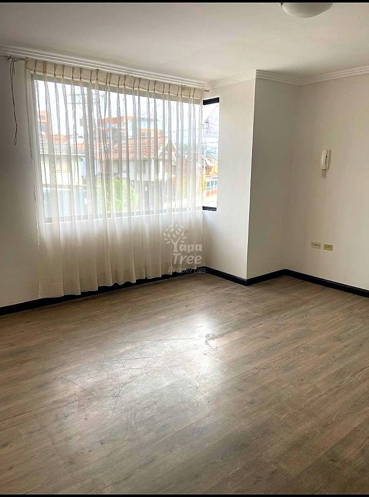 Empty bedroom with wood-look laminate flooring, white walls, large window with sheer curtains, and black base trim.