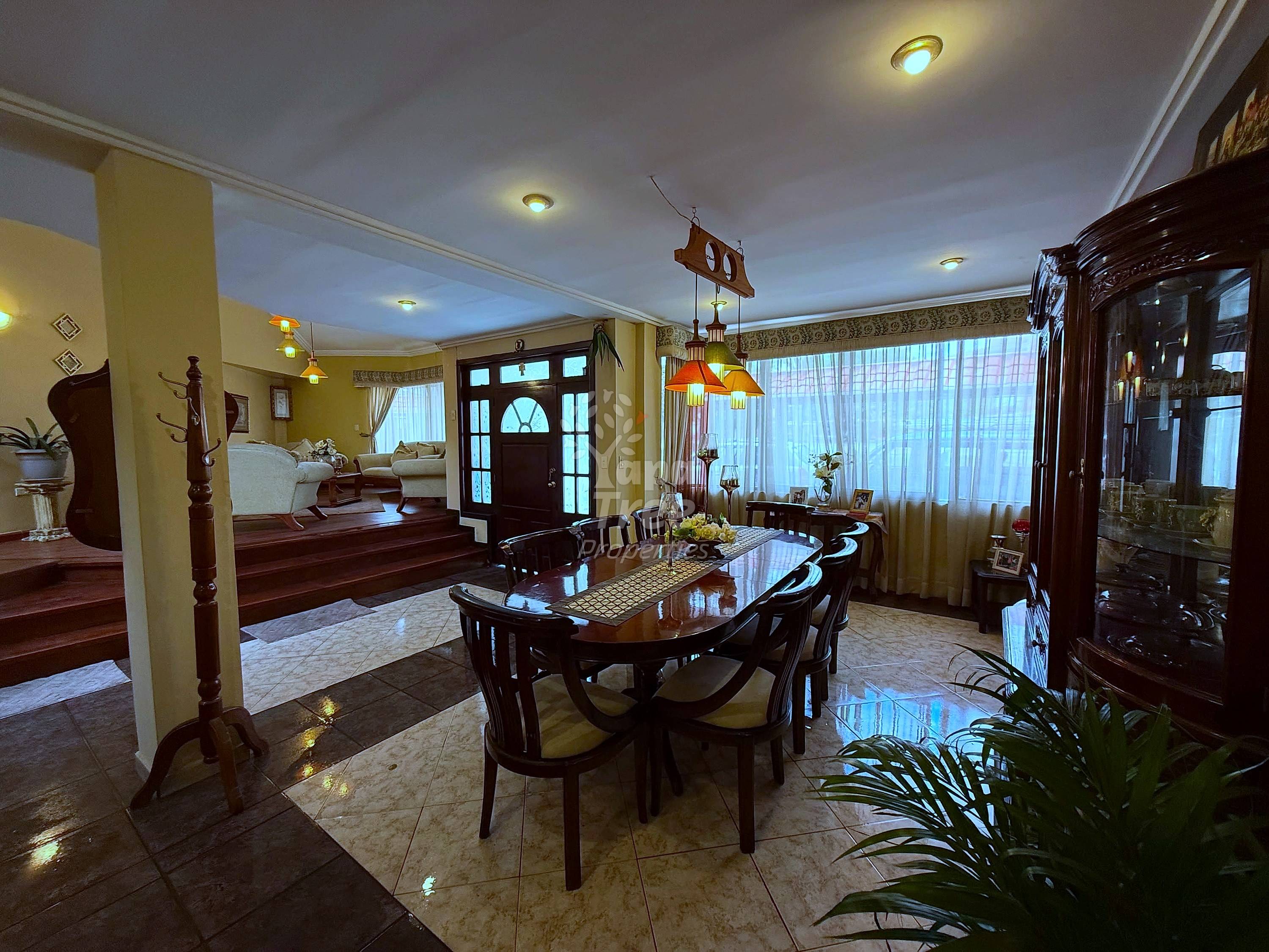 Dining room with dark wood table and chairs, cream walls, recessed ceiling lights, large windows with curtains, and dark wood display cabinet.