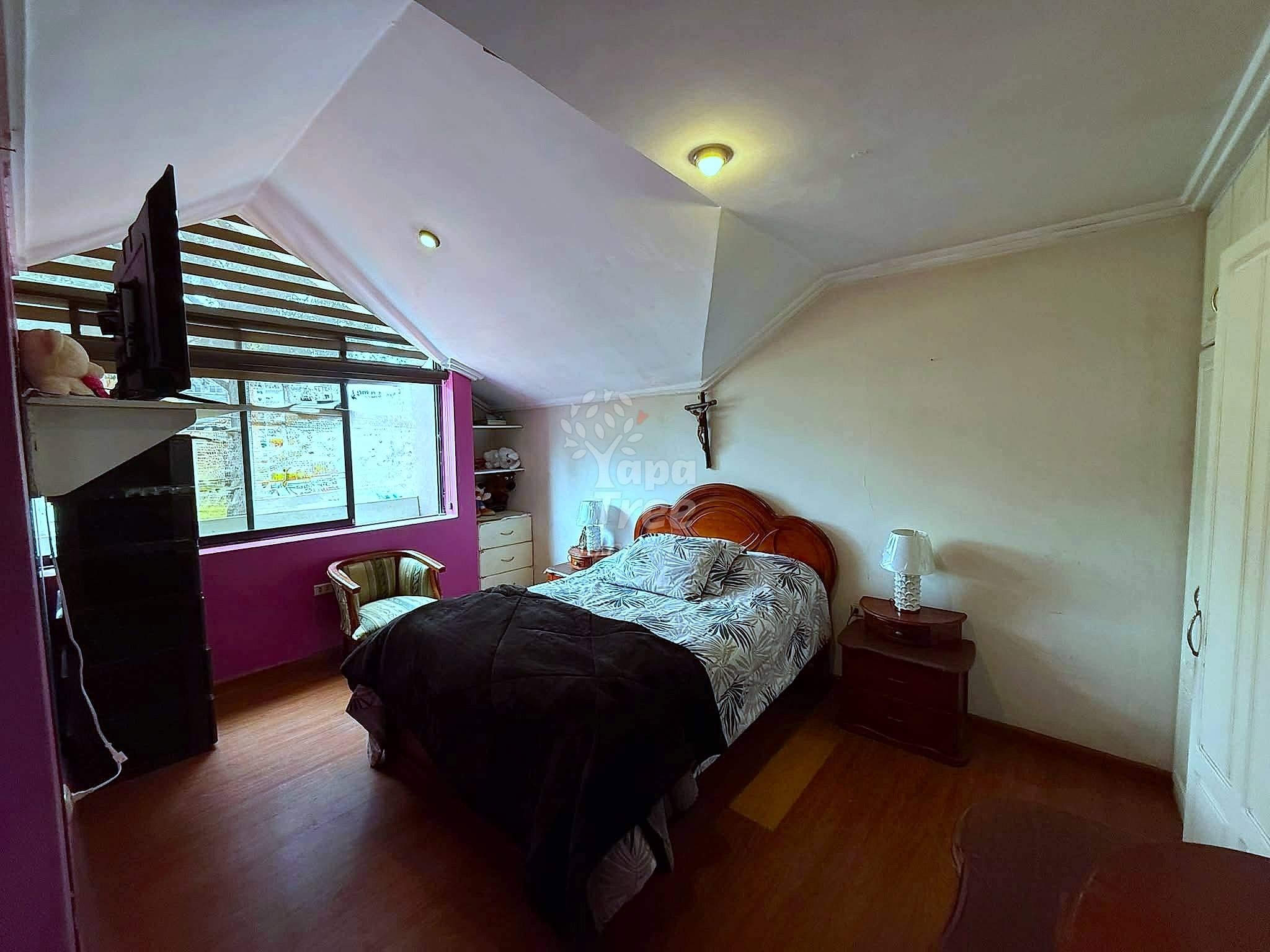 Bedroom with wooden bed frame, patterned bedding, nightstands with lamps, burgundy accent wall, angled ceiling with recessed lighting, and large window.