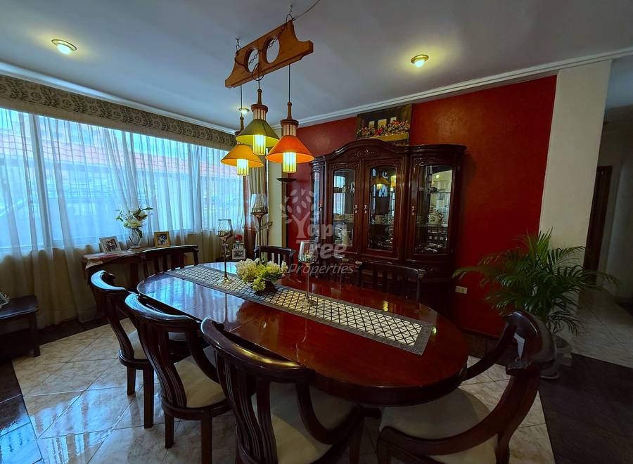 Dining room with dark wood table, matching chairs, orange pendant lights, burgundy accent wall, glass cabinet, and large windows with sheer curtains.