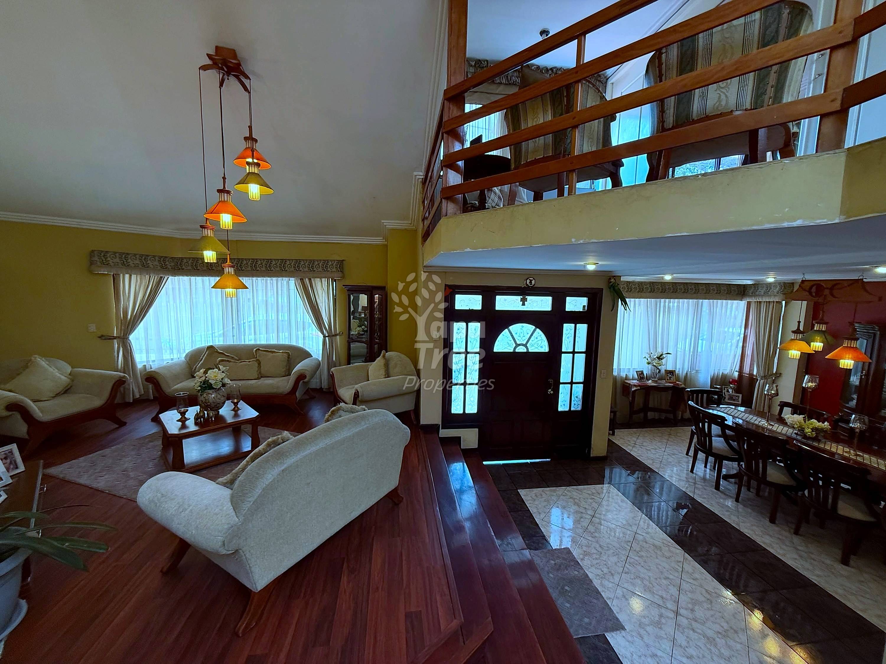 Open living room with cream-colored sofas, wooden floor, yellow walls, pendant lights, and dark entry door with transom window.