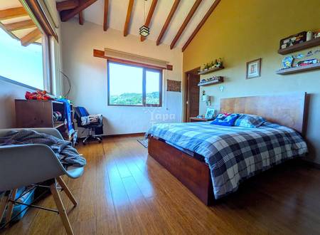 Bedroom with wooden bed frame, plaid bedding, wooden beams, large window with mountain views, wood flooring, desk, and wall shelving.