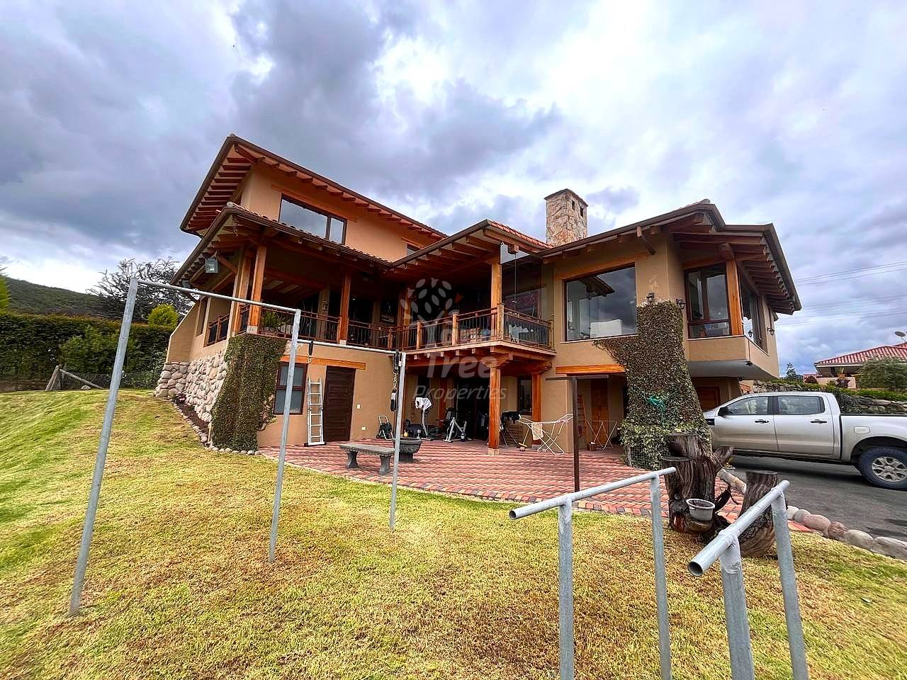 Two-story residential house with beige stucco walls, wooden beams, brick patio, and wraparound deck overlooking green lawn and mountains.