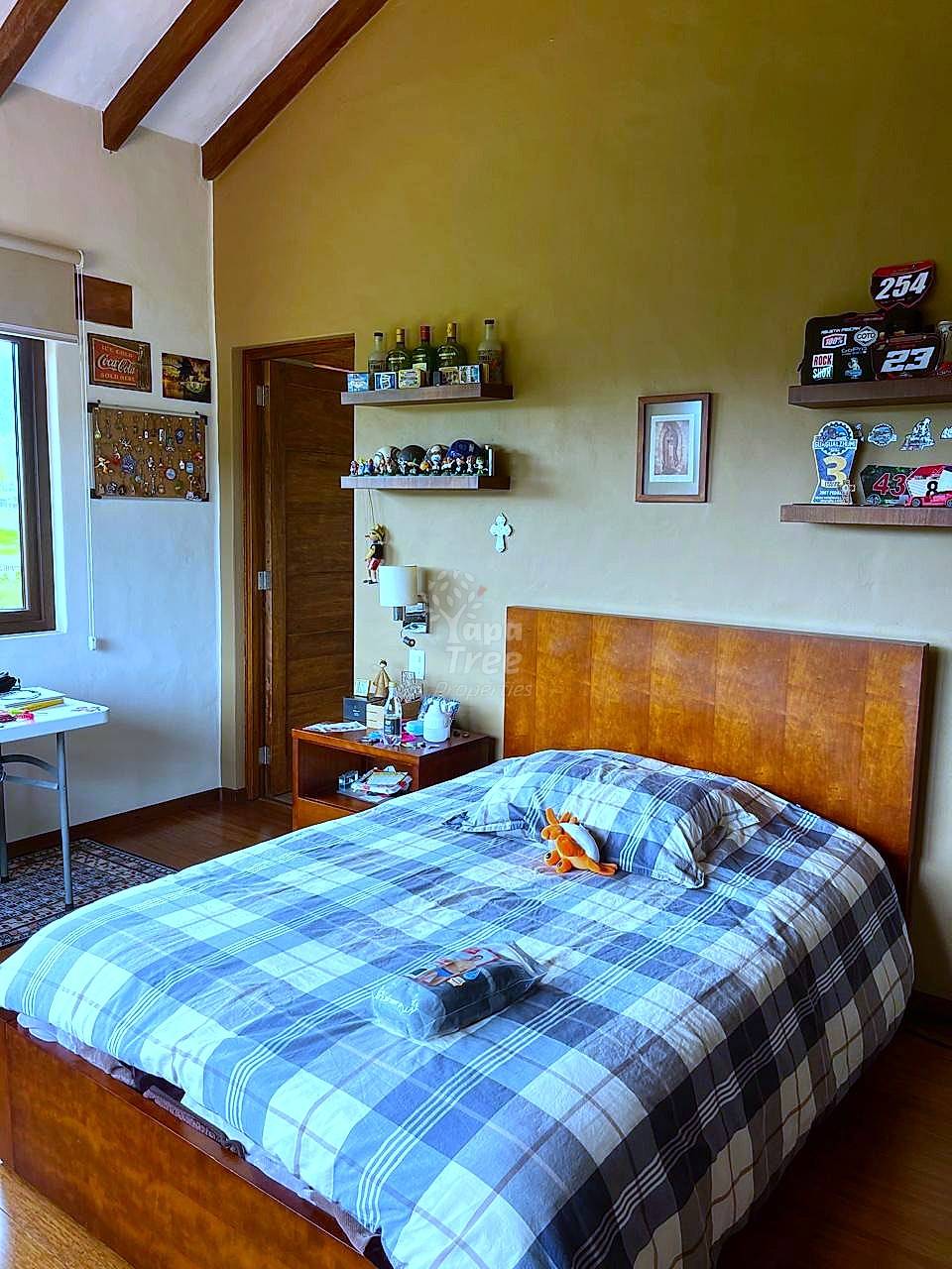 Bedroom with wooden bed frame, plaid bedding, exposed ceiling beams, yellow walls, wooden shelves, and desk area with natural light.