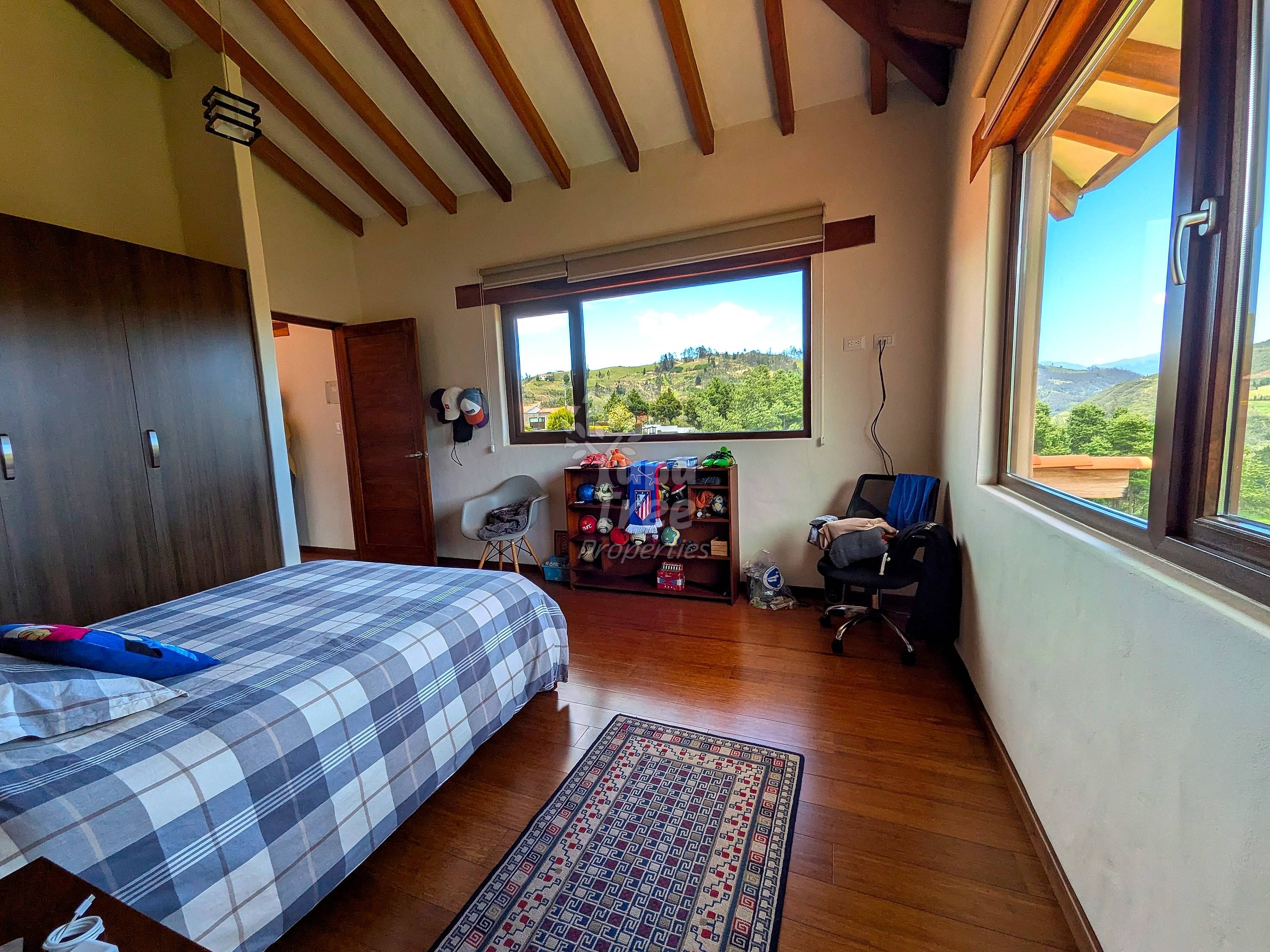 Bedroom with wooden beams, queen bed with checkered bedding, wooden floor, large windows with mountain views, wooden shelf unit, and desk chair.