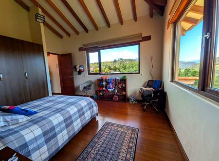 Bedroom with wooden beams, queen bed with checkered bedding, wooden floor, large windows with mountain views, wooden shelf unit, and desk chair.