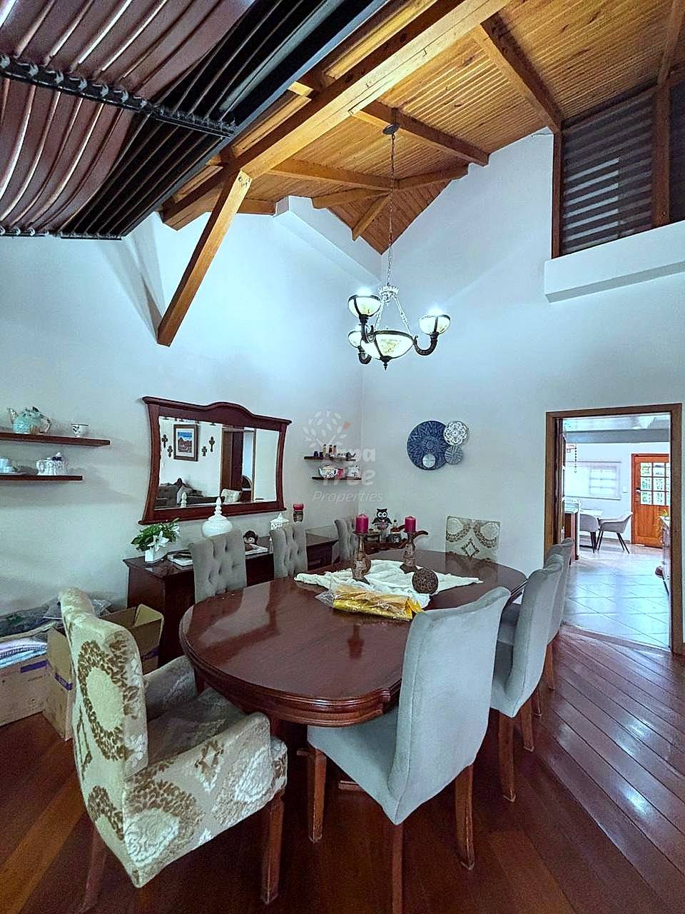 Dining room with exposed wooden beam ceiling, dark wood table, upholstered chairs, chandelier, and white walls with shelving.