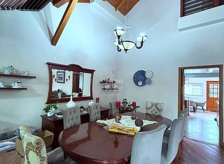 Dining room with exposed wooden beam ceiling, dark wood table, upholstered chairs, chandelier, and white walls with shelving.