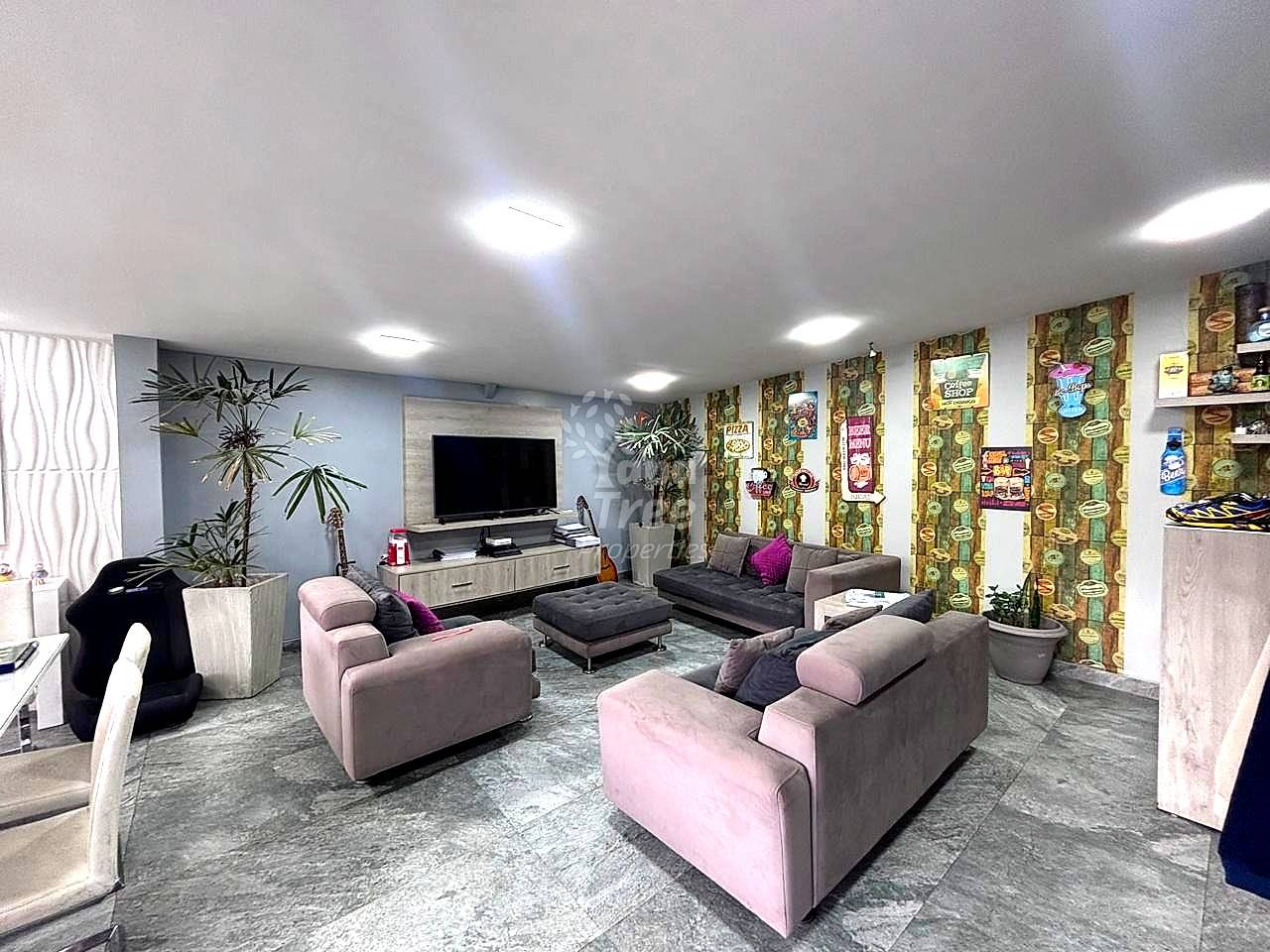 Large living room with pink and gray upholstered seating, flat-screen TV on wooden media console, potted plants, patterned wallpaper accent walls, LED ceiling lights, and gray tile flooring.
