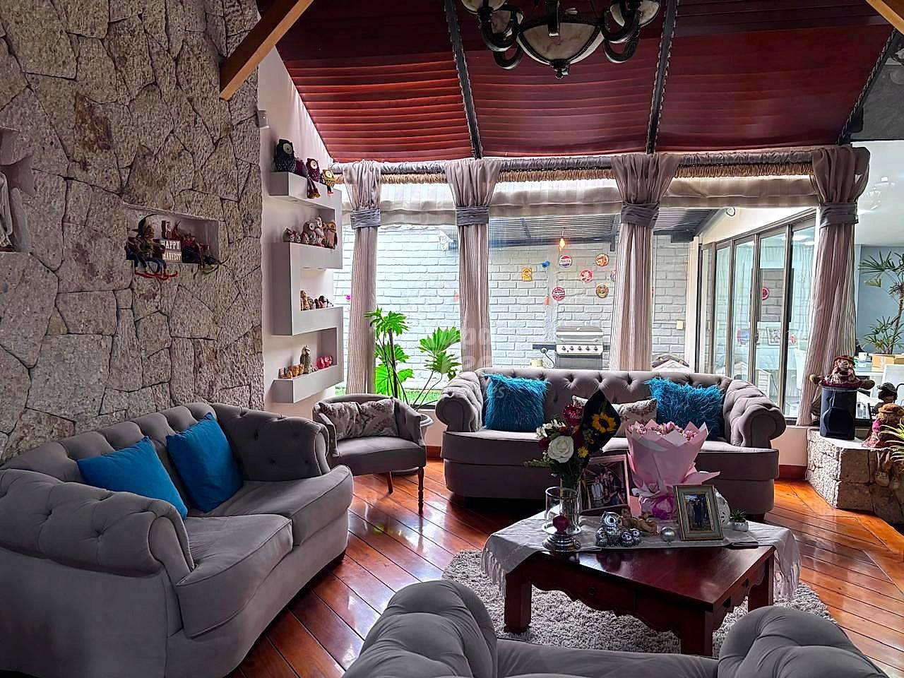 Living room with gray upholstered sofas, blue pillows, dark wood coffee table, wooden floors, high ceiling with red beams, and large windows with curtains.