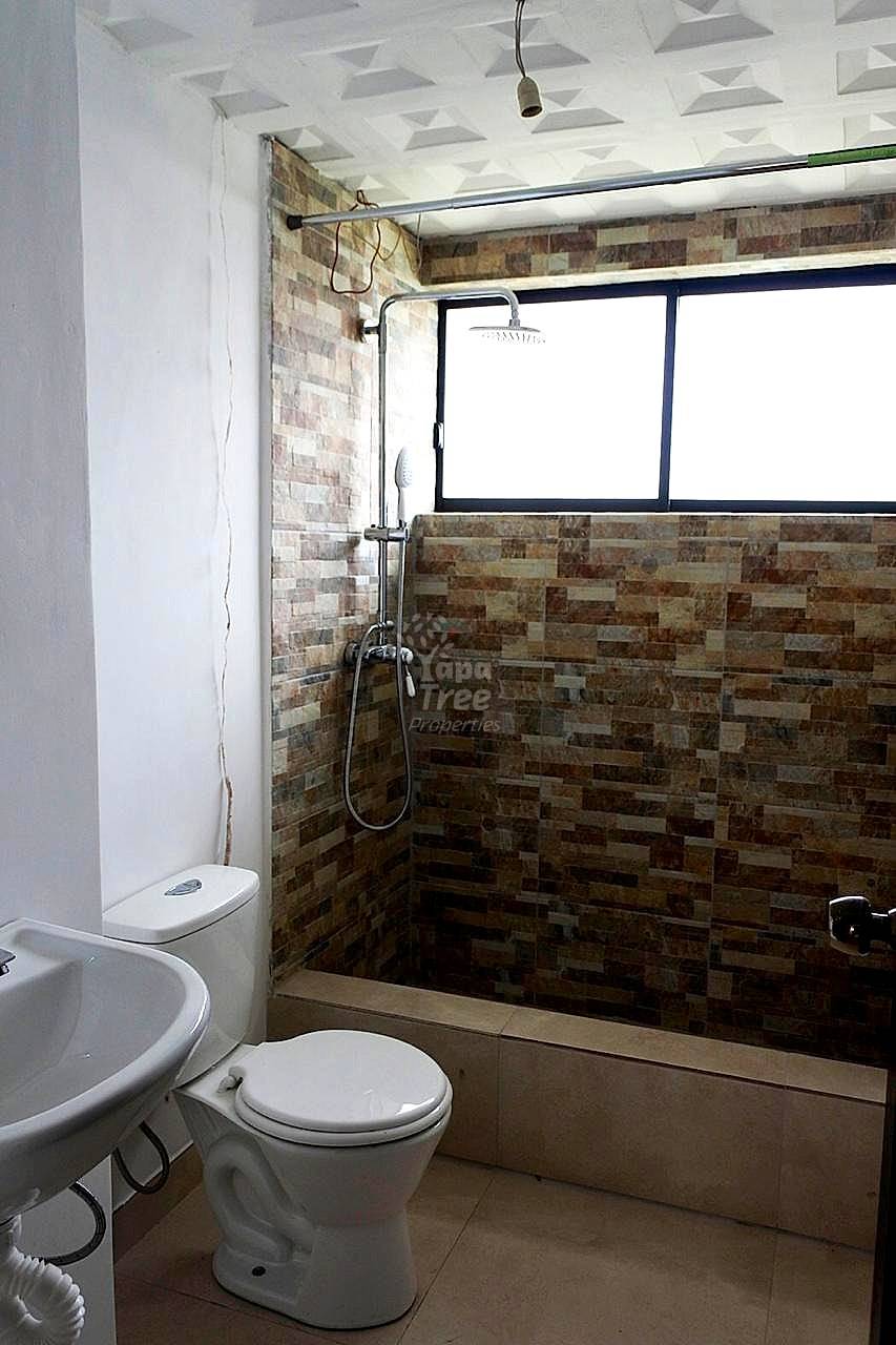Bathroom with white toilet, brown brick tile walls, glass shower enclosure with rainfall showerhead, and window with dark frame.