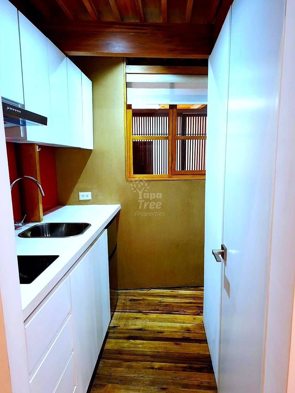Narrow galley cocina with white cabinetry, black stainless steel sink, wooden beams, yellow walls, and window with wooden shutters.