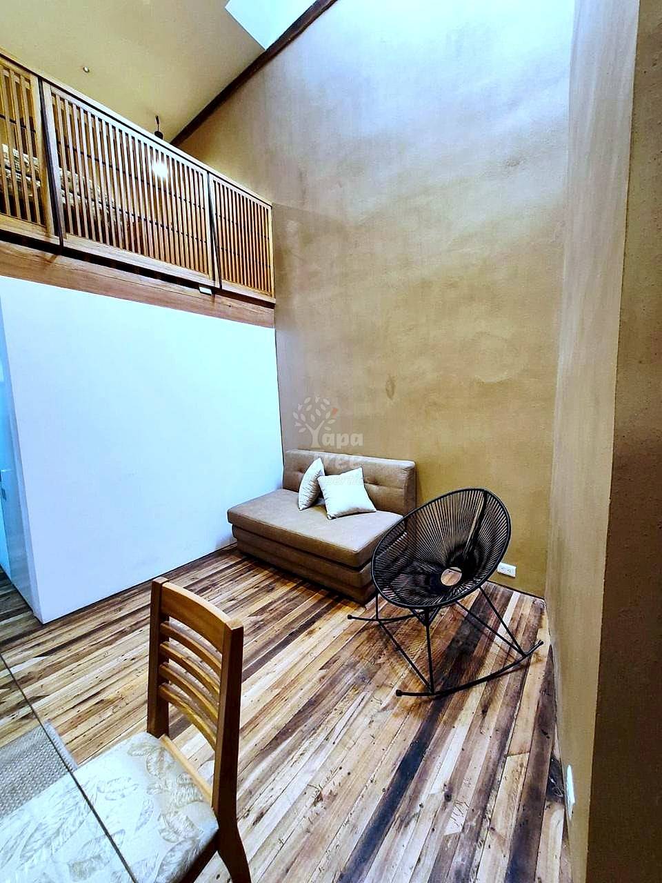 Loft-style room with high ceilings, mezzanine railing, tan walls, wood flooring, tan sofa with pillows, black chair, and wooden side table.
