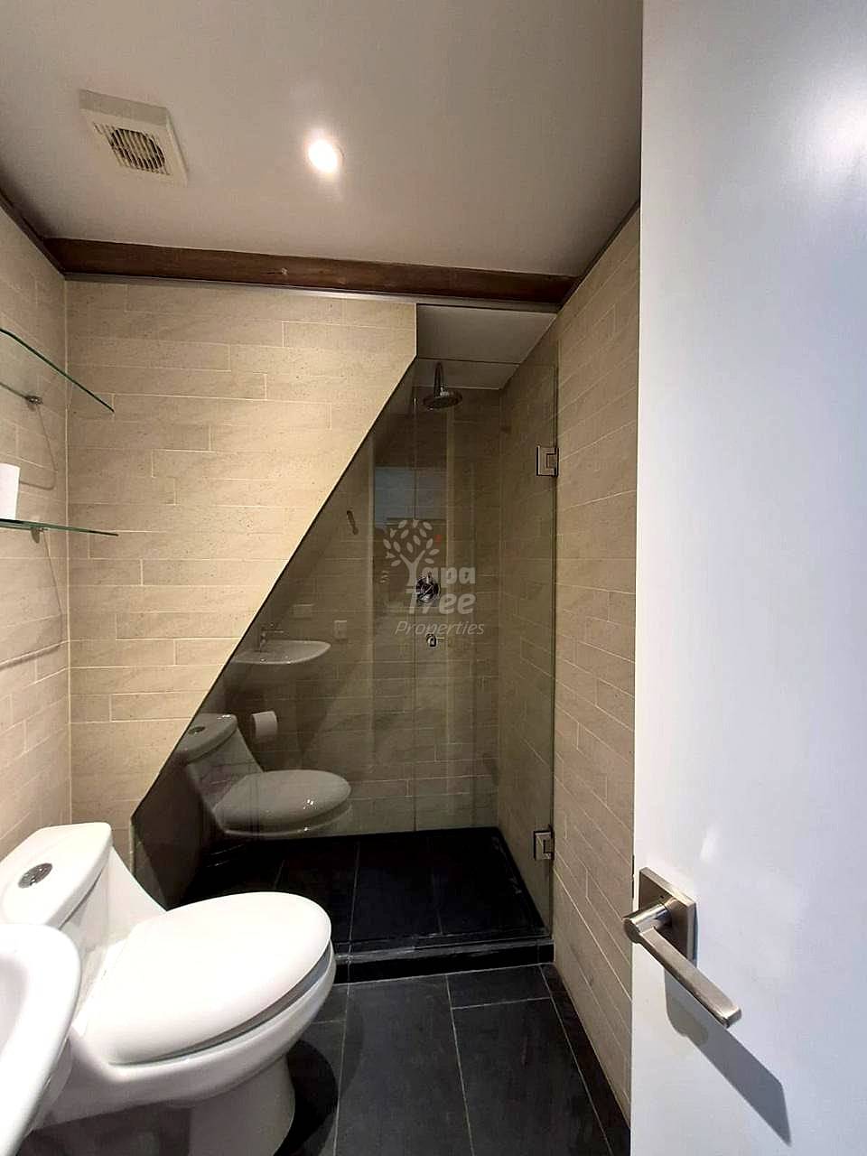 Bathroom with white toilet, beige tile walls, black tile floor, glass-enclosed shower stall, recessed ceiling light, and wood trim.
