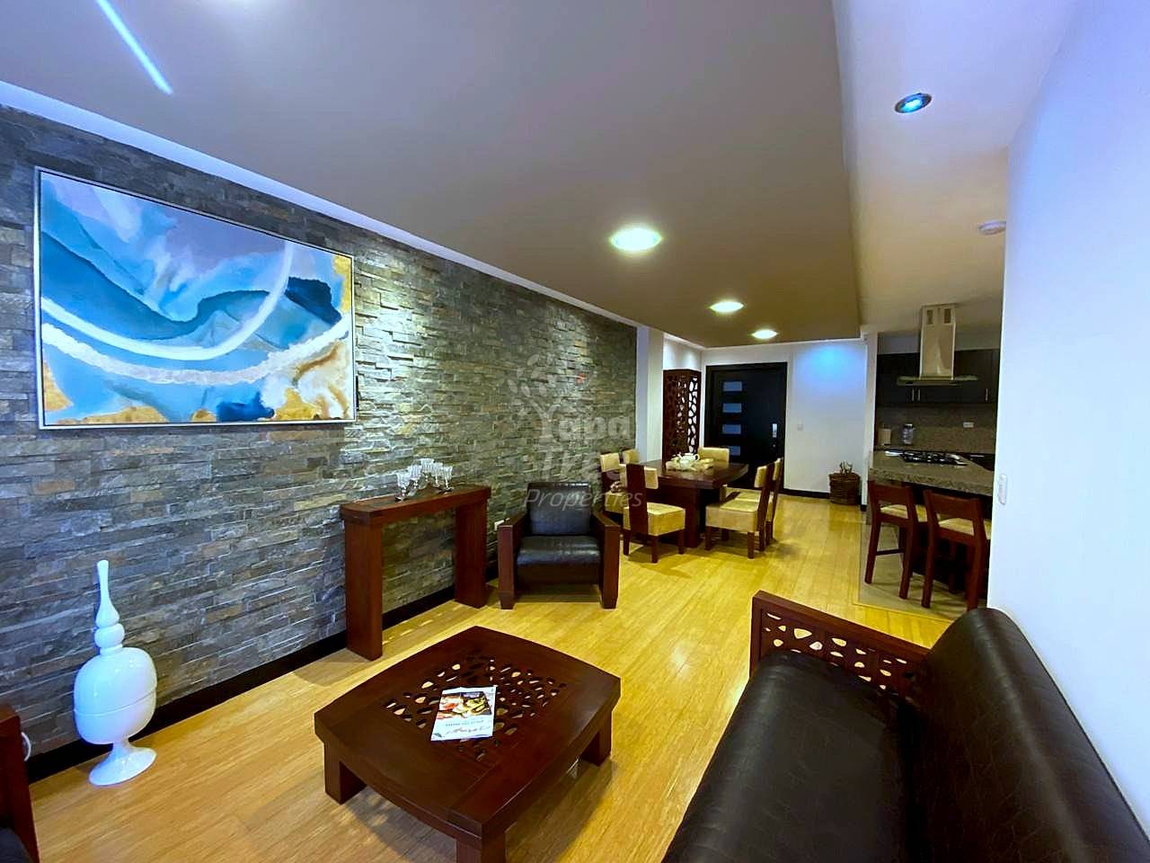 Open-plan living area with wood flooring, stone accent wall, recessed lighting, black leather sofa, wooden furniture, and modern kitchen visible in background