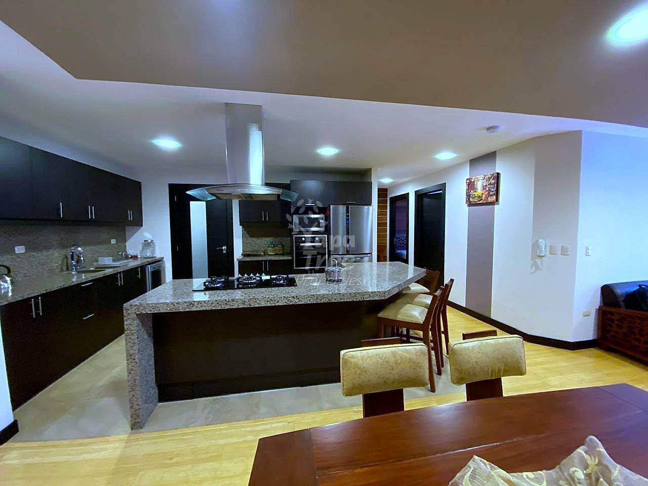 Open-plan cocina with black cabinets, granite countertops, stainless steel appliances, and island seating for three, with dining table and hardwood floors.