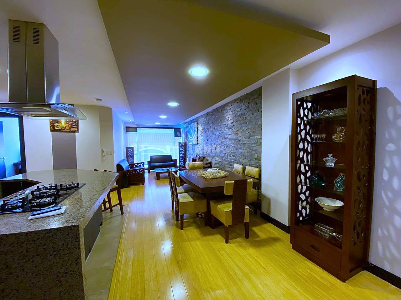 Open-concept departamento with kitchen featuring gas cooktop and stainless steel hood, dining area with wooden table, living room seating, wooden flooring, recessed ceiling lights, and decorative dark wood display cabinet.