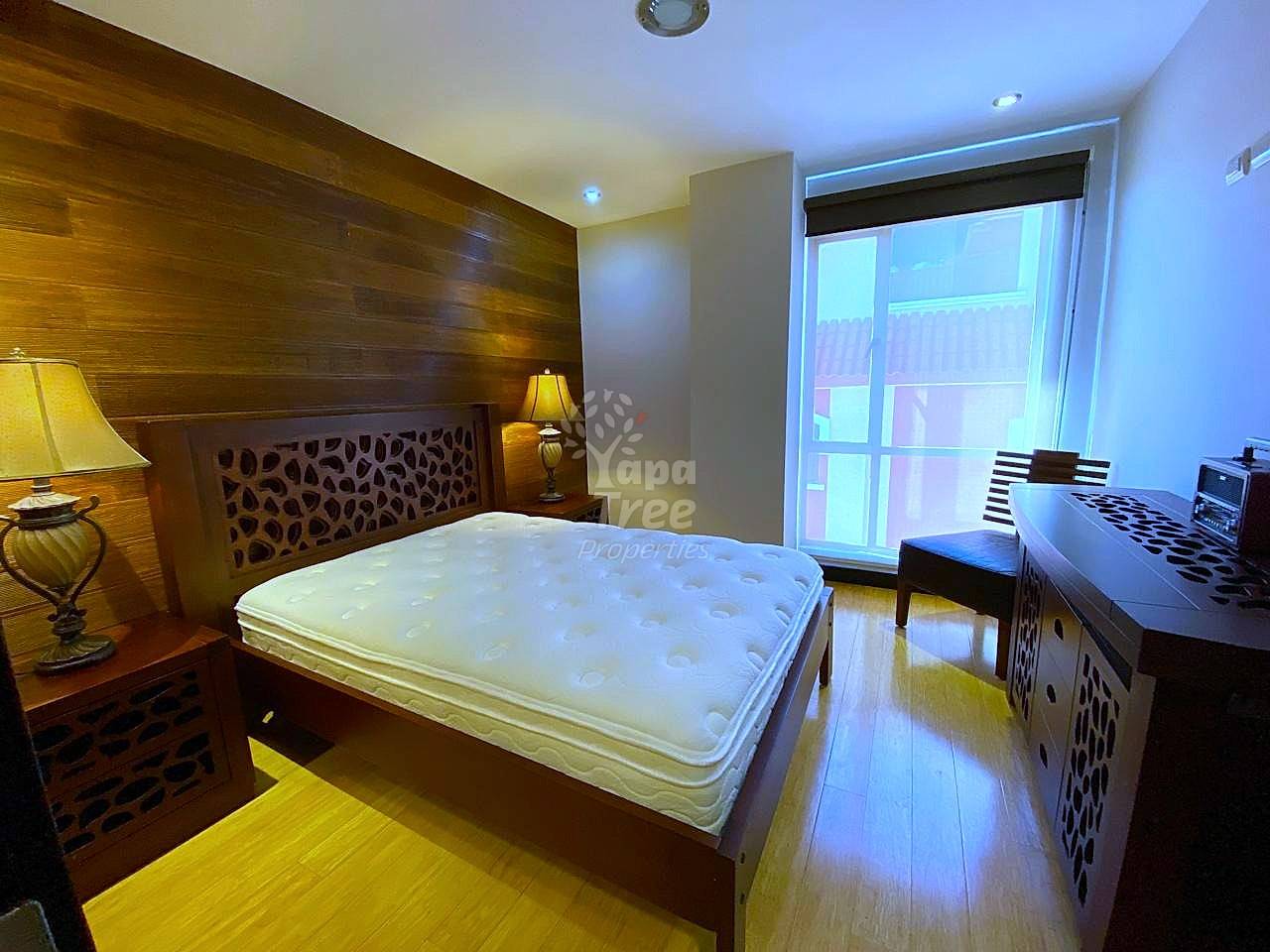 Bedroom with wood-frame bed, white mattress, patterned headboard, bedside lamp, wooden nightstand, large window with view, wooden flooring, and desk area with keyboard.