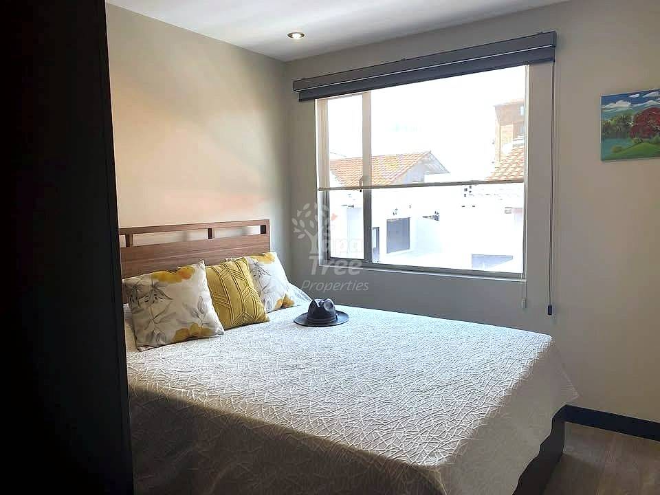 Bedroom with double bed, wood headboard, yellow decorative pillows, large window with gray frame overlooking rooftops, recessed ceiling light.